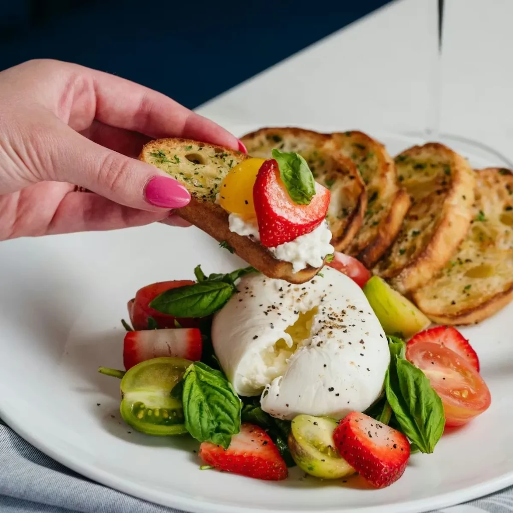 Creamy burrata, strawberries, tomatoes, basil; hand holding toasted bread.