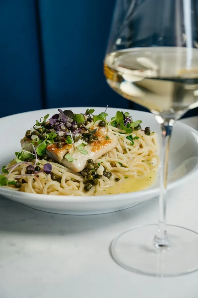 Pasta and fish dish with capers, microgreens, and white wine glass.