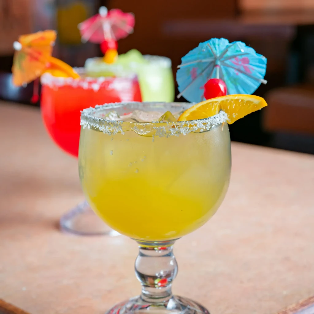 Yellow cocktail with salt rim, umbrella, cherry, and two blurred cocktails.