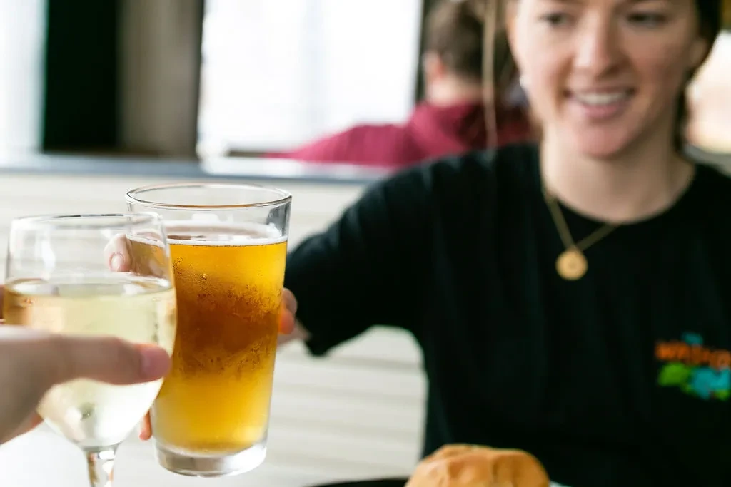 Two hands toast a wine glass and a beer glass in a restaurant.