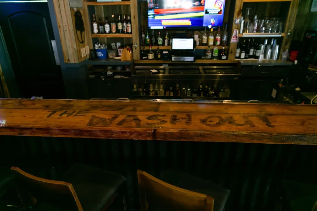 Dimly lit bar with 'THE WASH OUT' counter, liquor shelves, sports TV.