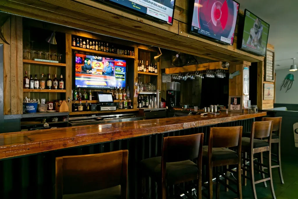 Rustic bar with wooden counter, liquor bottles, and multiple sports TVs.