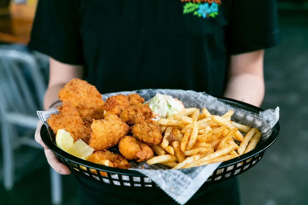 Black basket of fried fish, shrimp, fries, and coleslaw, held by person.