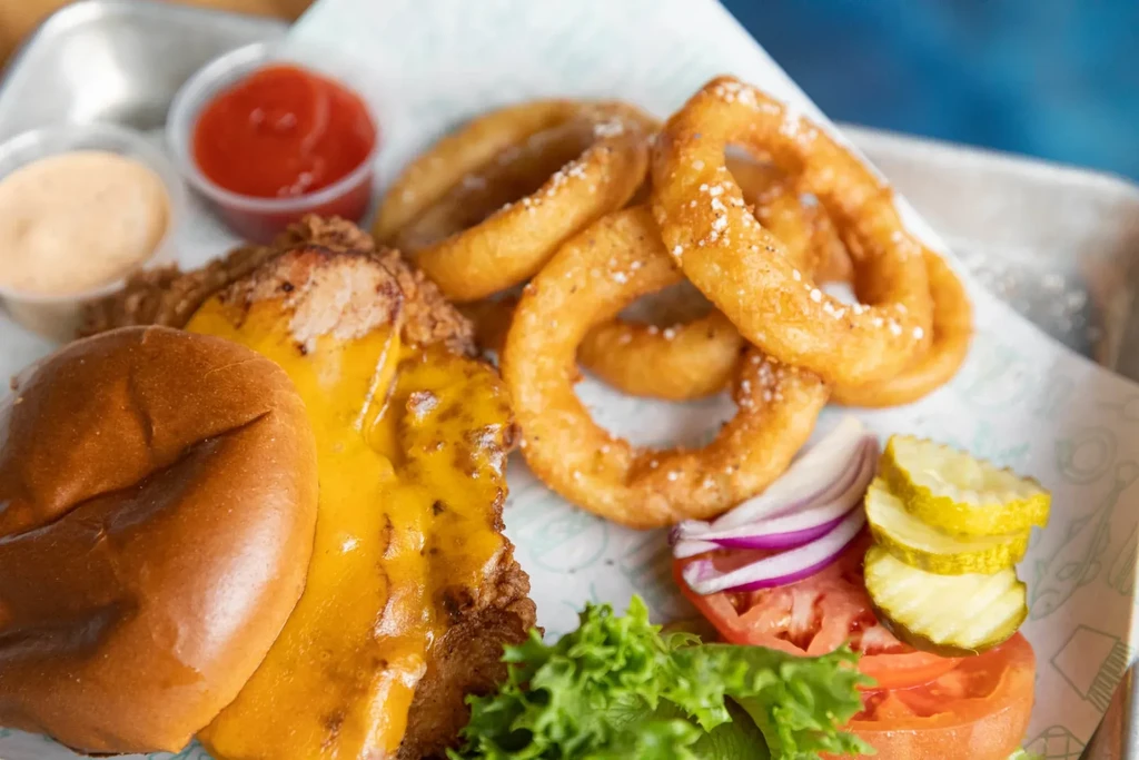 Fried chicken sandwich with melted cheese, onion rings, and garnish on tray.