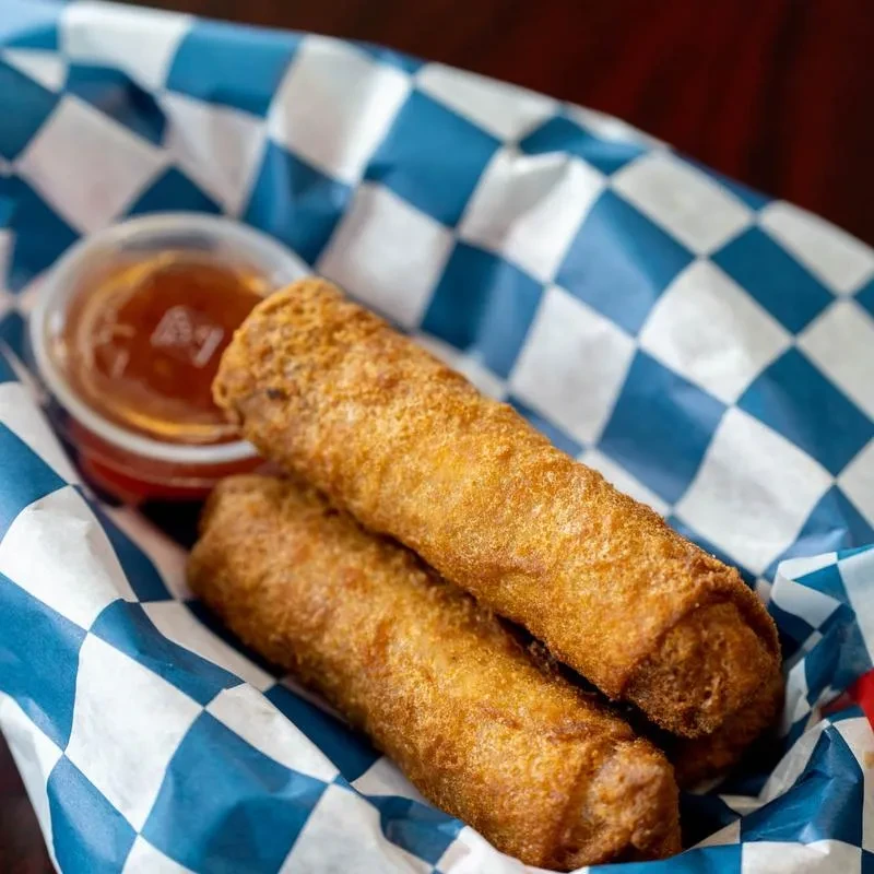 Crispy fried rolls and amber dipping sauce on checkered paper.