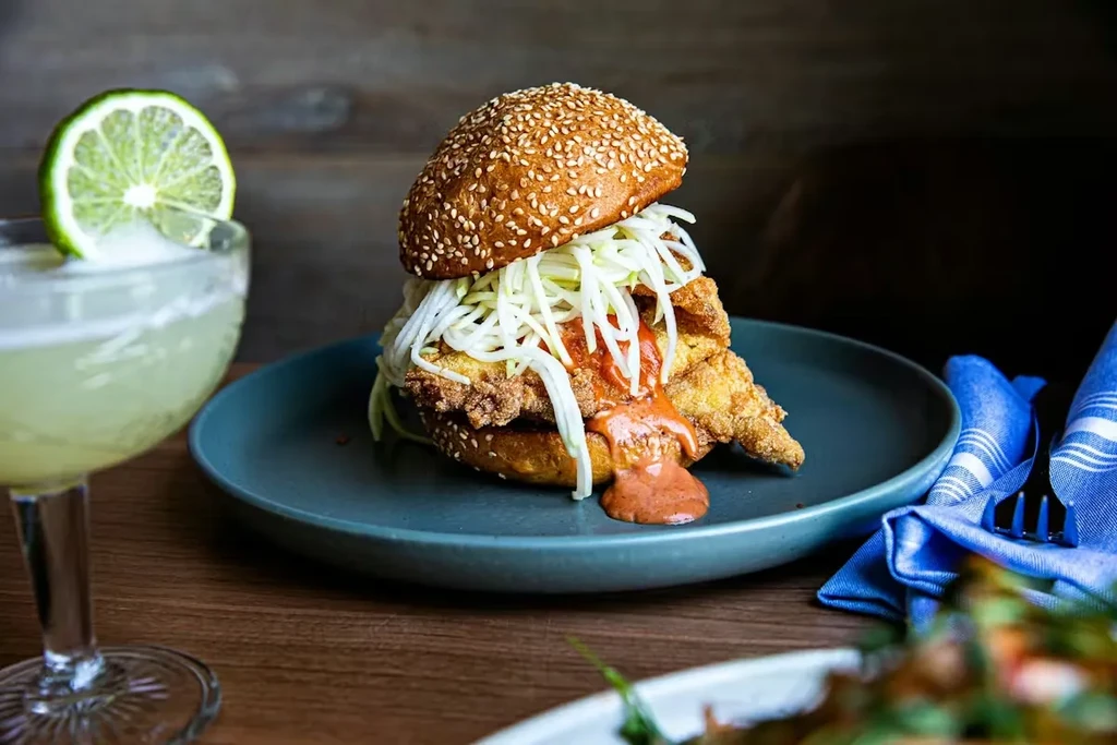 Fried chicken sandwich with coleslaw and sauce, beside a lime cocktail.