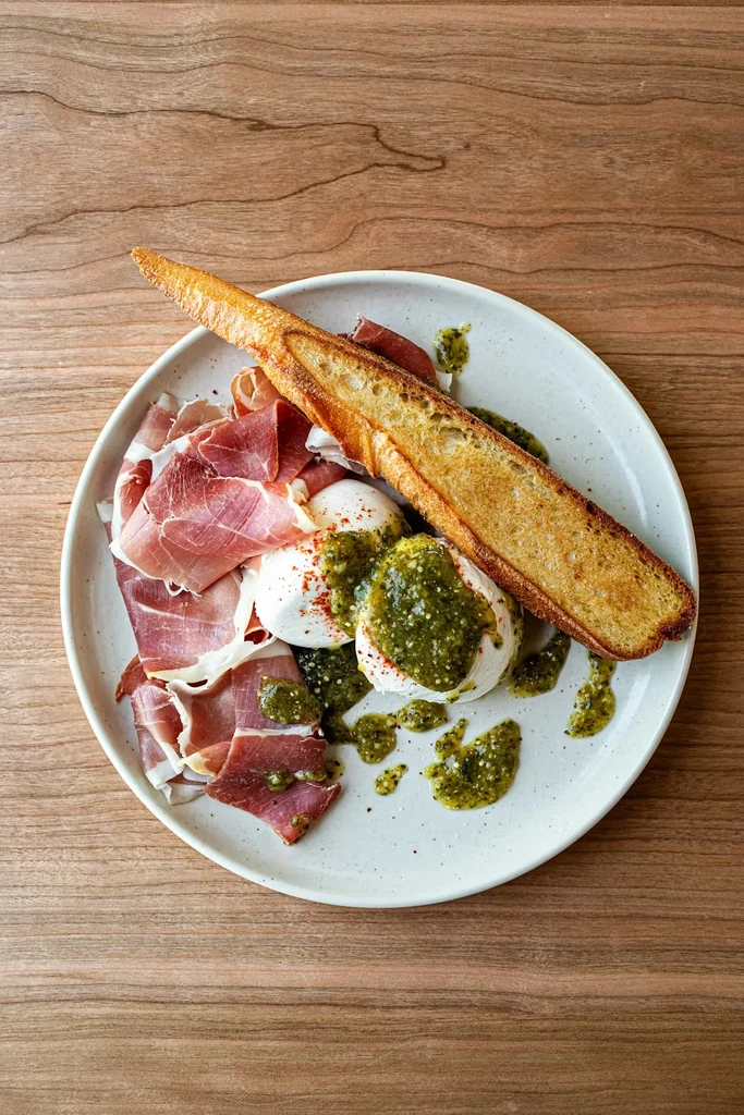 Burrata, prosciutto, pesto, and toasted baguette on a speckled plate.