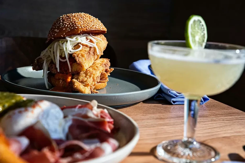 Fried chicken sandwich, cocktail, and charcuterie platter on a wooden table.