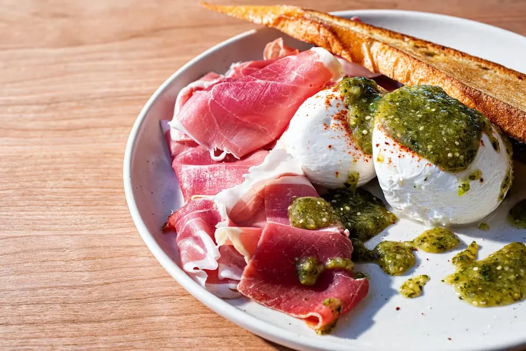 Prosciutto, burrata with green pesto, red flakes, and bread on plate.