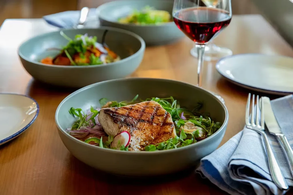 Grilled fish/poultry with greens, wine, cutlery on a wooden table.