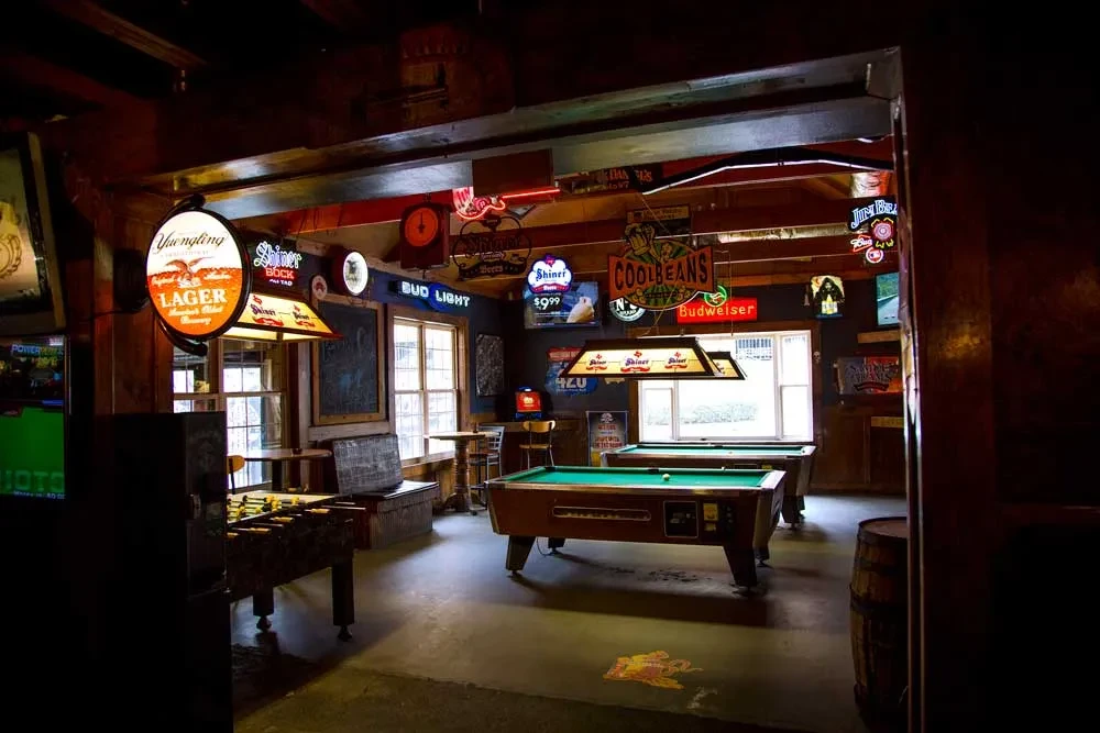 Rustic pub interior with two pool tables, foosball, and glowing beer signs.