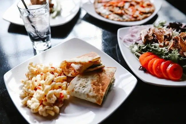 Restaurant table with macaroni salad, grilled meat salad, wraps, and drink.