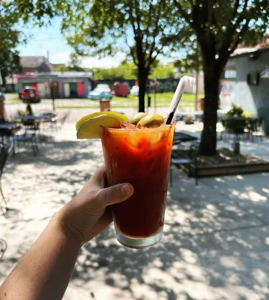 Hand holding Bloody Mary with lime and olives on sunny patio.