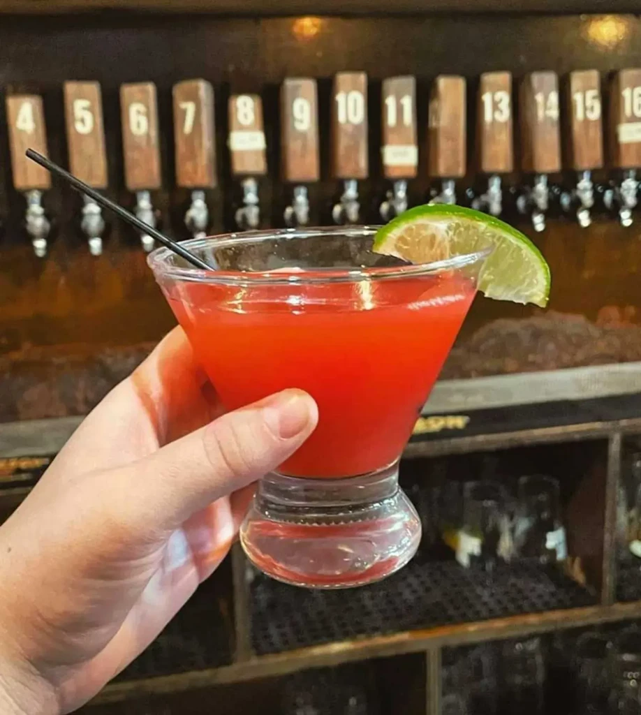 Hand holding red cocktail with lime and straw in a bar.