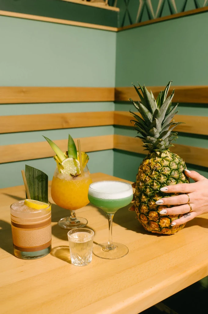 Three cocktails, shot glass, hand holding pineapple on wooden table.