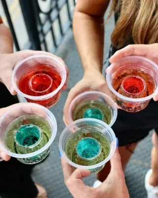 Multiple hands hold five red and teal bomb shots in plastic cups.