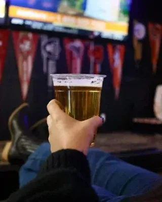 Hand holds foamy beer in plastic cup at a dimly lit bar.