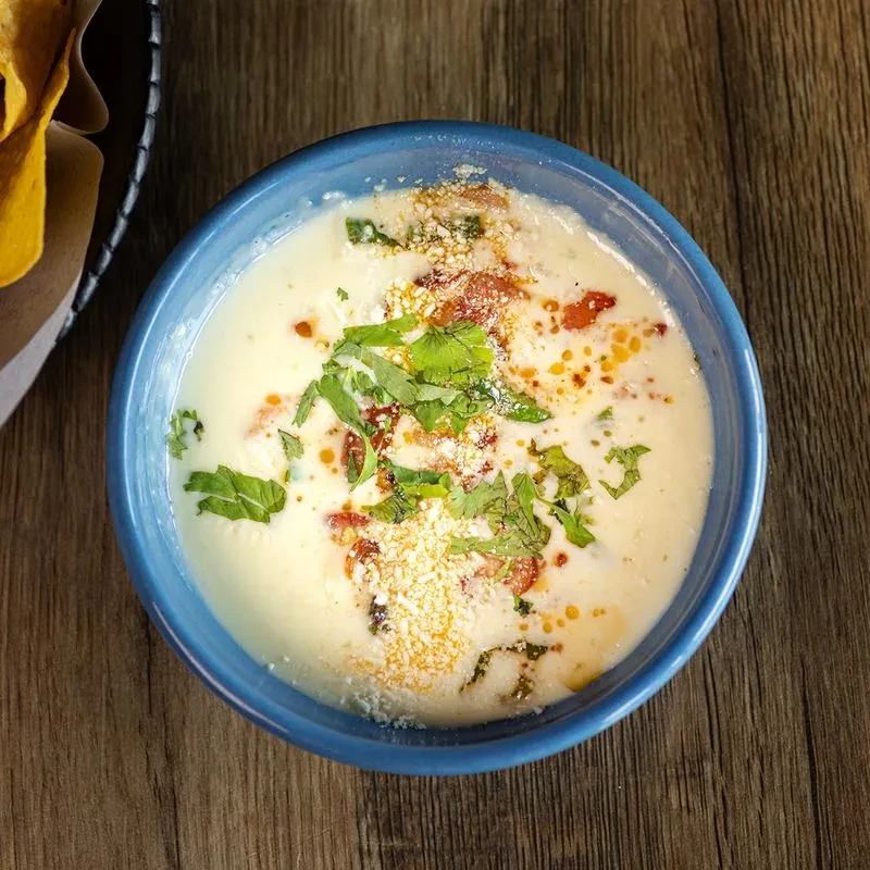Queso dip in blue bowl with cilantro, chips, on wooden table.