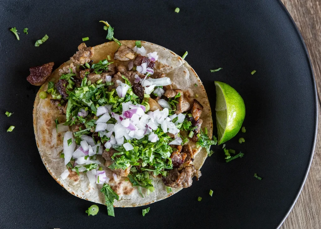 Single street taco with meat, cilantro, onions, lime on a black plate.