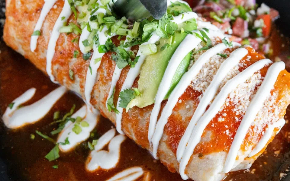 Overhead burrito with reddish sauce, avocado, crema, cheese, and herbs.