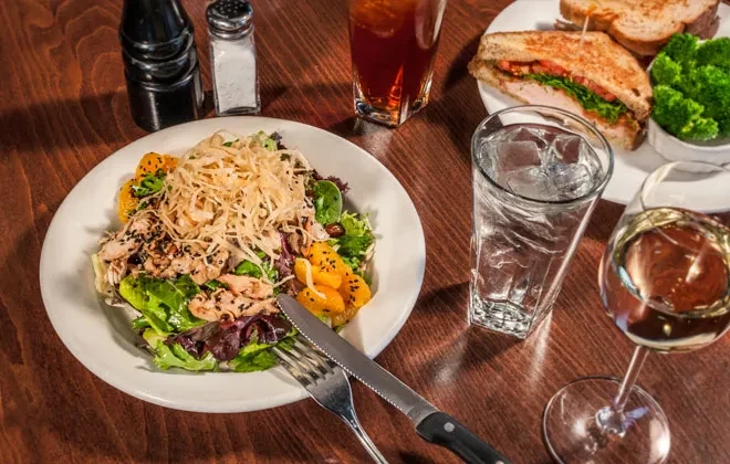 Overhead: vibrant salad, sandwich, drinks on a wooden dining table.