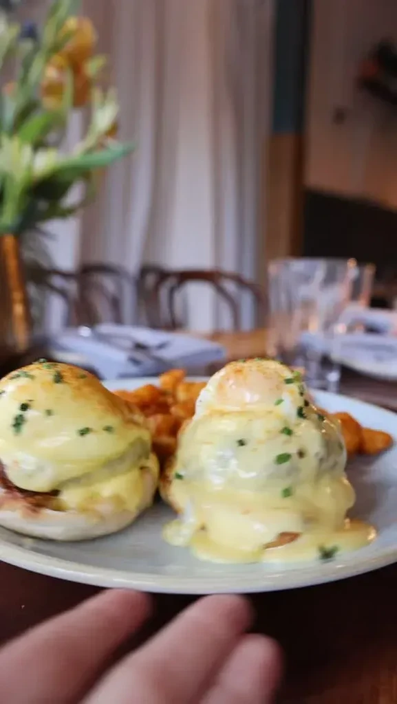 Close-up of Eggs Benedict with hollandaise sauce, hash browns, and blurred hand.