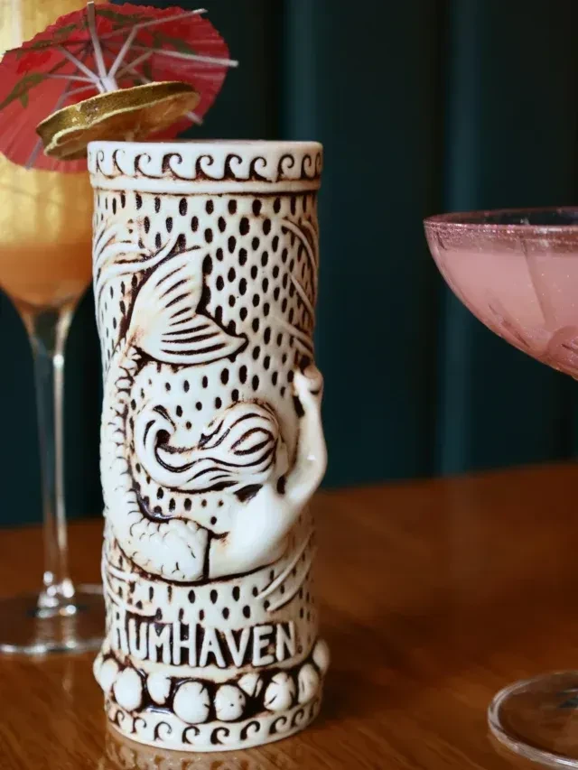 Garnished tiki mug and pink cocktail on wood, blurred dark background.