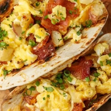 Two close-up breakfast tacos with eggs, potatoes, bacon, and chives.