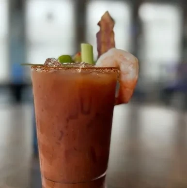 Reddish cocktail garnished with shrimp, bacon, celery, and olives.