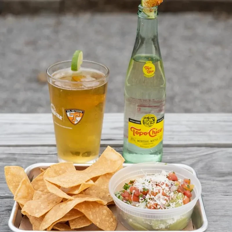 Beer, Topo Chico, tortilla chips, and guacamole on a wooden outdoor table.