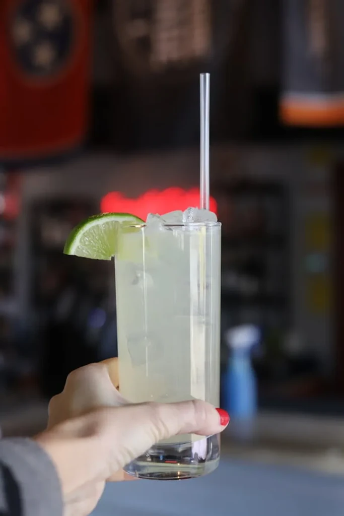 Hand holding glass of pale green drink with lime wedge and ice.