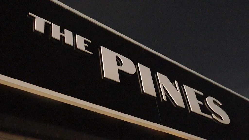 Metallic 'THE PINES' sign with raised silver letters on dark panel.