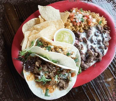 Red plate with two soft tacos, chips, rice, beans, and salsa.