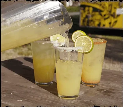 Silver pitcher pours lime-garnished margaritas into rimmed glasses outdoors.