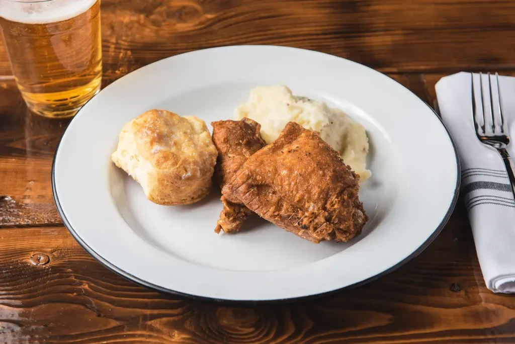 Fried chicken, mashed potatoes, biscuit, and beer on a rustic wooden table.