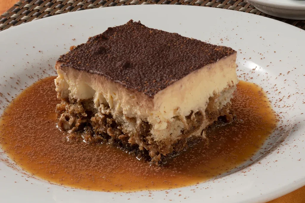 Layered tiramisu slice, cocoa-dusted, with sauce on a plate.