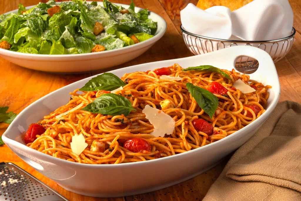 Spaghetti, Caesar salad, and bread rolls on a rustic wooden table.