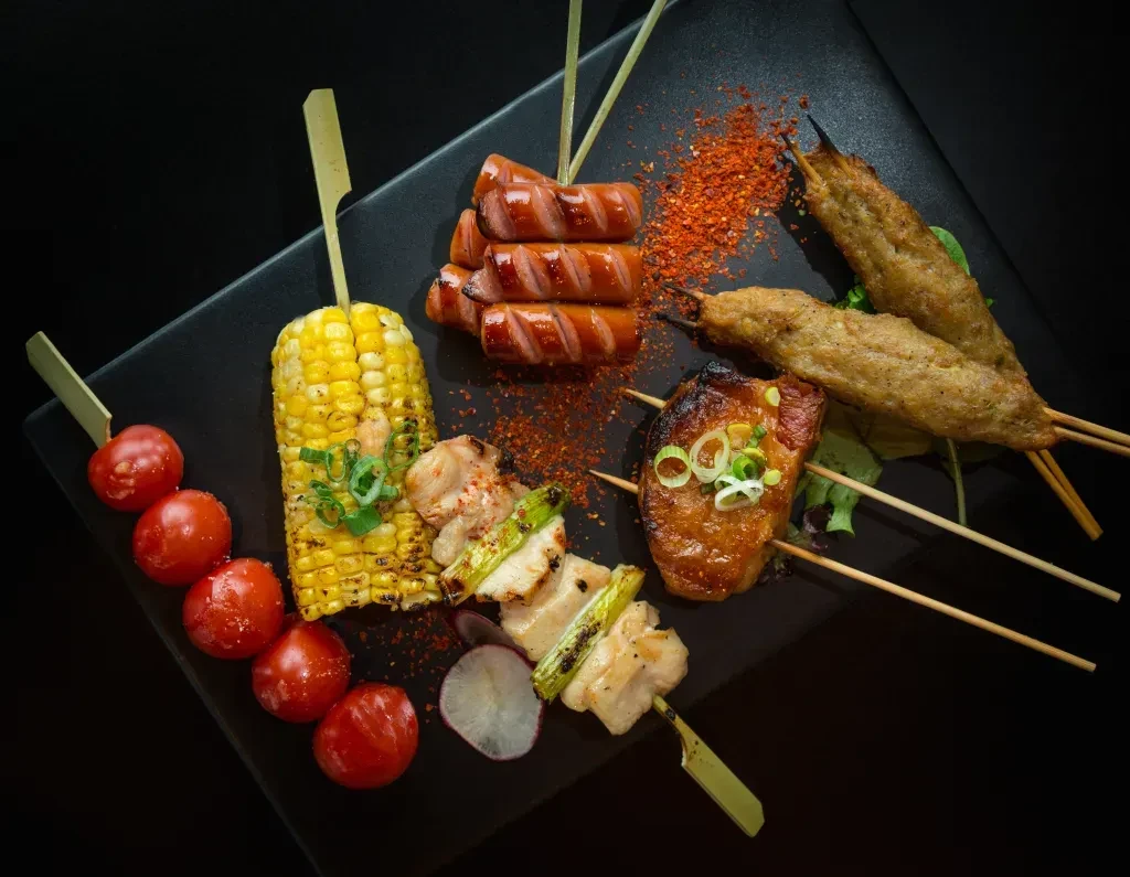 Assorted grilled skewers, sausages, and corn on a dark plate.