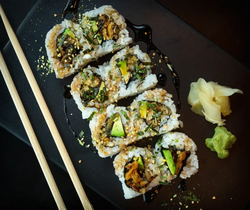 Six inside-out sushi rolls, avocado, ginger, wasabi on a dark plate.