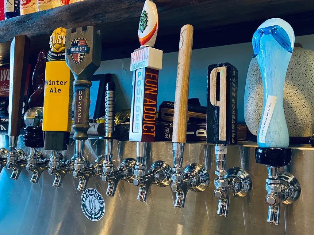Eight unique beer tap handles on a shiny stainless steel bar.