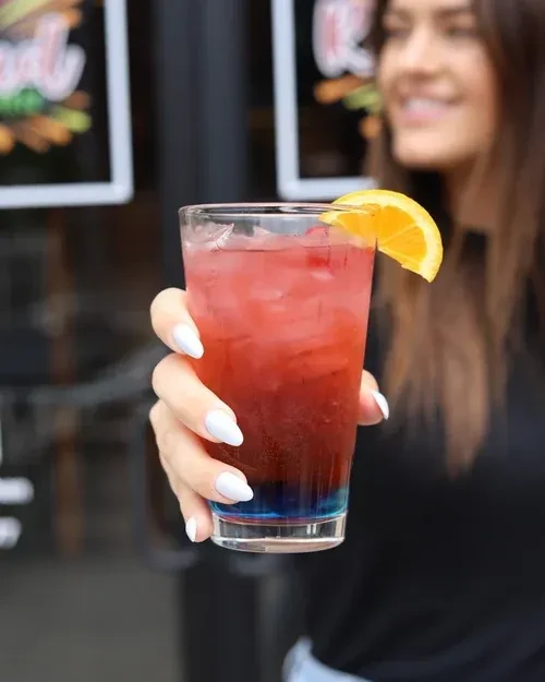 Woman's hand holds layered blue, red, pink cocktail with orange slice.
