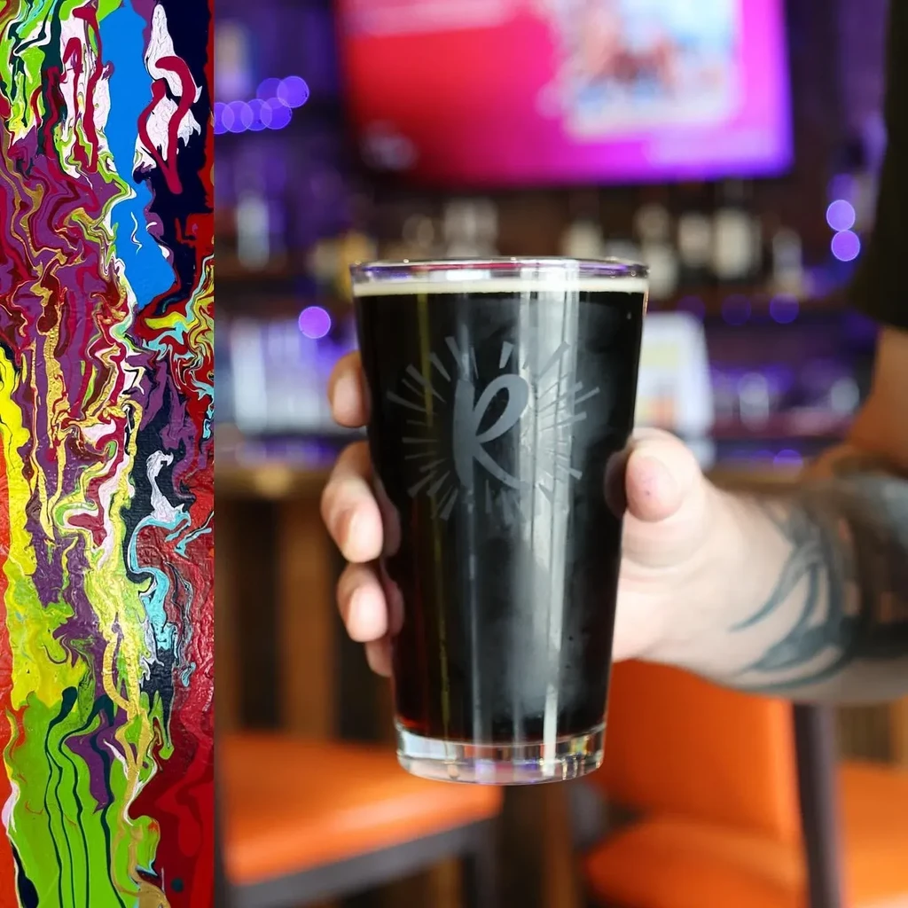 Tattooed hand holding dark beverage in an etched glass.