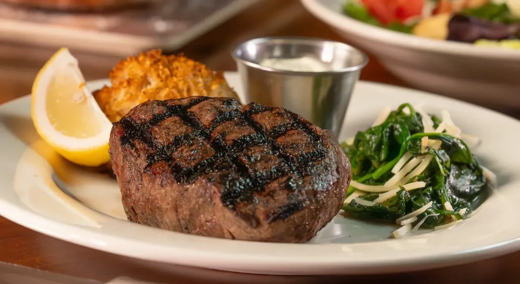 Grilled steak, lemon, spinach, and crispy fritter on a white plate.