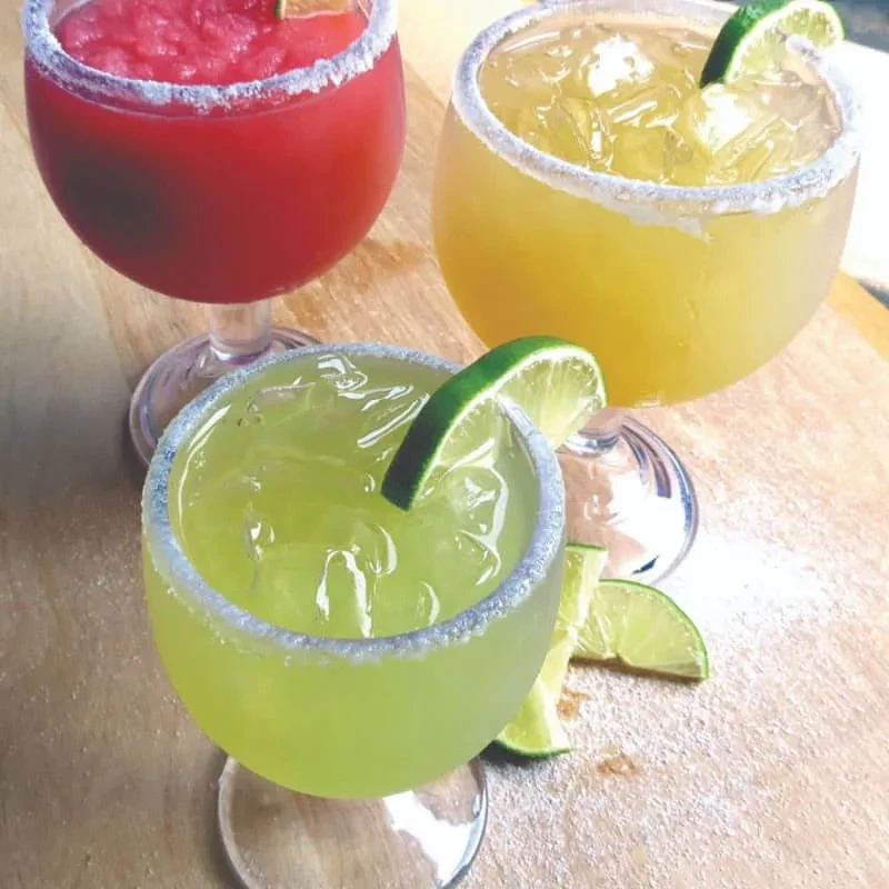 Three colorful cocktails with lime wedges and salted rims on wood.