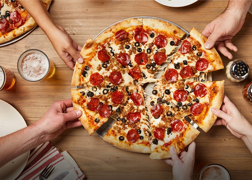 Hands reach for slices of pepperoni, olive, and sausage pizza with beer.