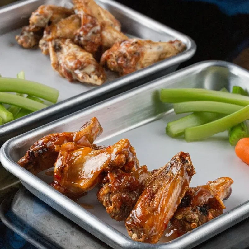 Two metal trays of sauced and dry-rub chicken wings with celery.