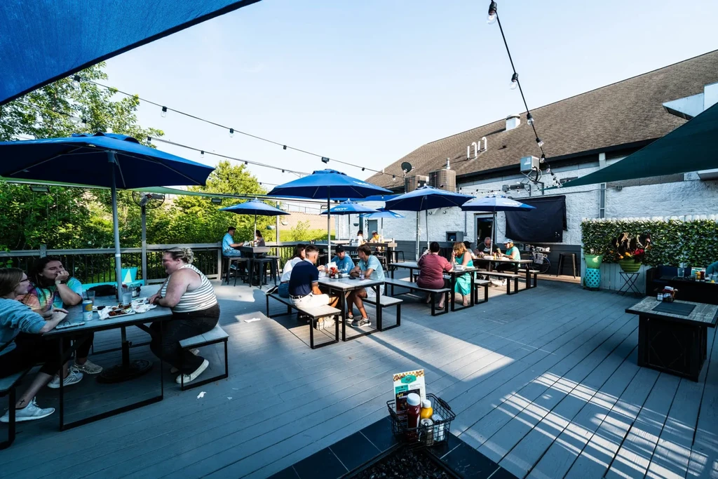 Sunny outdoor restaurant patio with people, picnic tables, and blue umbrellas.