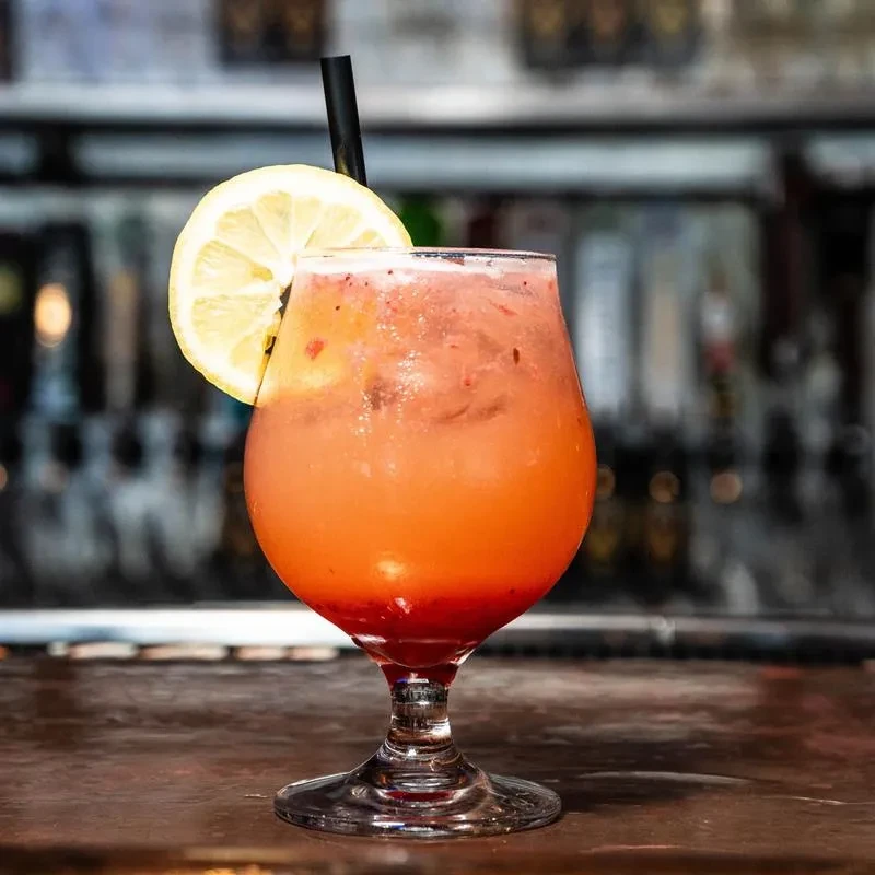 Reddish-orange cocktail with muddled fruit, lemon, and straw on a bar.