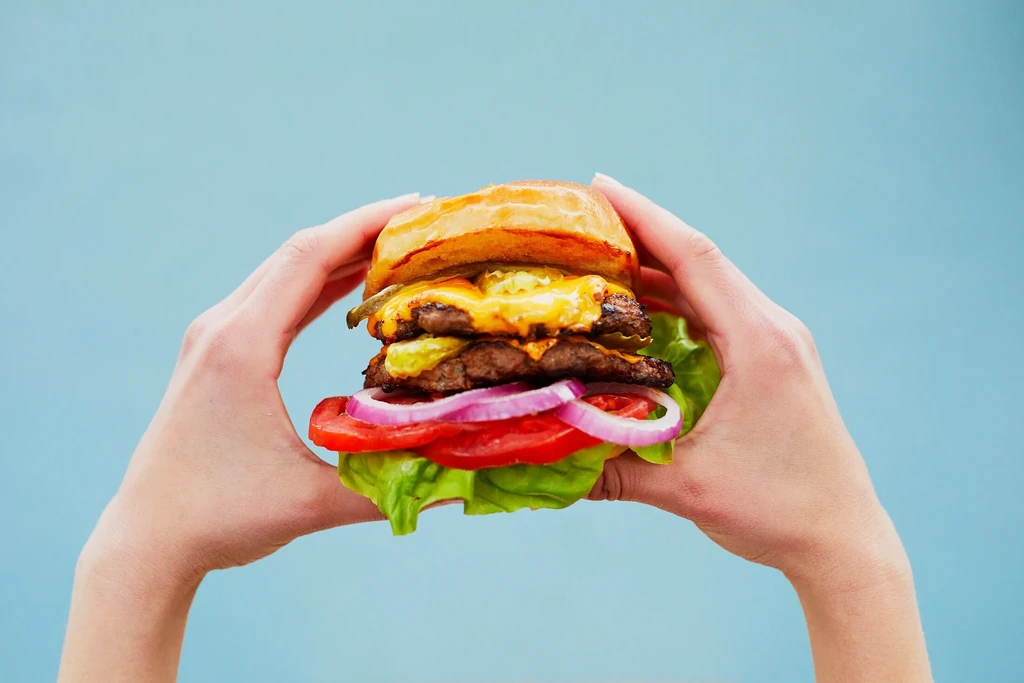 Well-lit double cheeseburger with cheese and vegetables, held by hands.