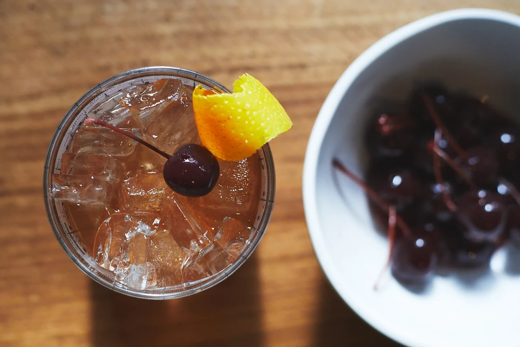 Overhead shot: amber cocktail, ice, cherry, citrus peel, and cherries.